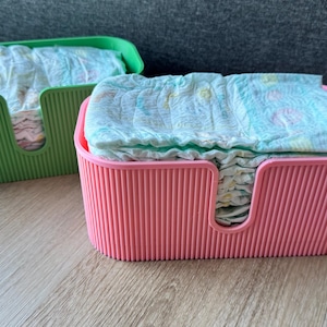 May include: Two rectangular storage bins with vertical ribbed textures, one in bright green and one in light pink, each featuring a U-shaped cutout on the front side and filled with a stack of folded disposable diapers with light blue patterns.