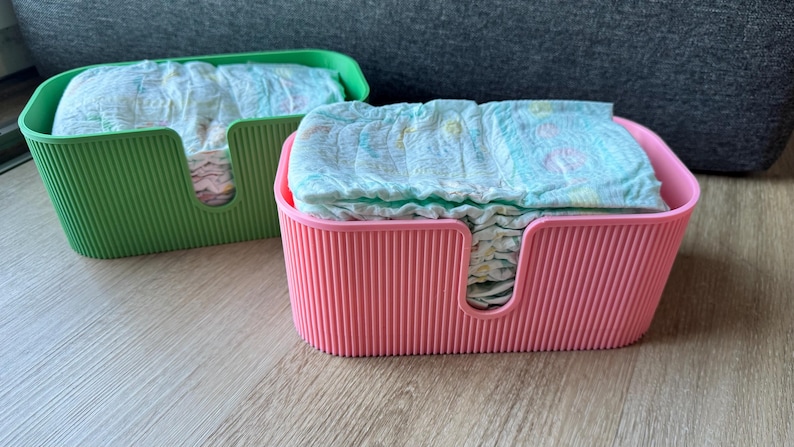 May include: Two rectangular storage bins with vertical ribbed textures, one in bright green and one in light pink, each featuring a U-shaped cutout on the front side and filled with a stack of folded disposable diapers with light blue patterns.