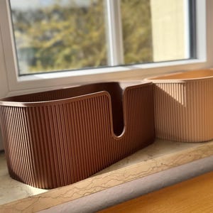 May include: Two oval-shaped storage bins with a vertical ribbed texture. The front bin is dark brown and features a U-shaped cutout on the front panel, while the partially visible bin behind it is a light beige color with the same ribbed design.