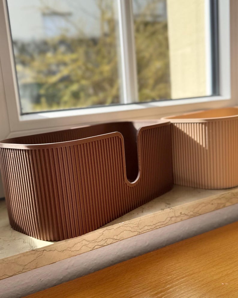 May include: Two oval-shaped storage bins with a vertical ribbed texture. The front bin is dark brown and features a U-shaped cutout on the front panel, while the partially visible bin behind it is a light beige color with the same ribbed design.
