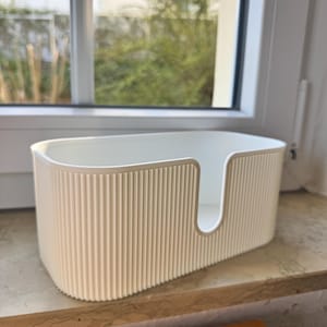 May include: An off-white, oval-shaped storage bin featuring a vertical ribbed texture on the exterior walls. The container has a smooth, rounded rim and a U-shaped cutout on one of the long sides, providing an open access point for items.
