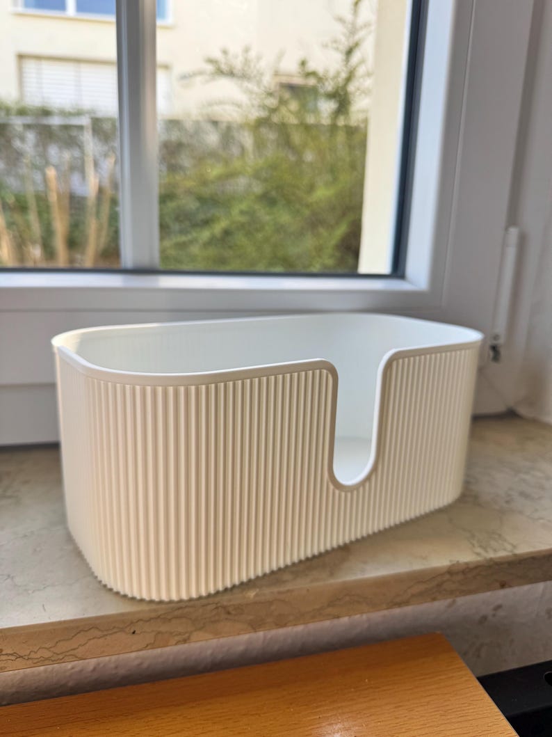 May include: An off-white, oval-shaped storage bin featuring a vertical ribbed texture on the exterior walls. The container has a smooth, rounded rim and a U-shaped cutout on one of the long sides, providing an open access point for items.