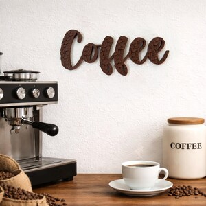 May include: A brown wooden "Coffee" sign hangs on a white wall above a coffee machine and a cup of coffee. The sign is in a cursive font with a textured design. Jars labeled "Coffee" and "Sugar" sit nearby, along with burlap sacks filled with coffee beans.