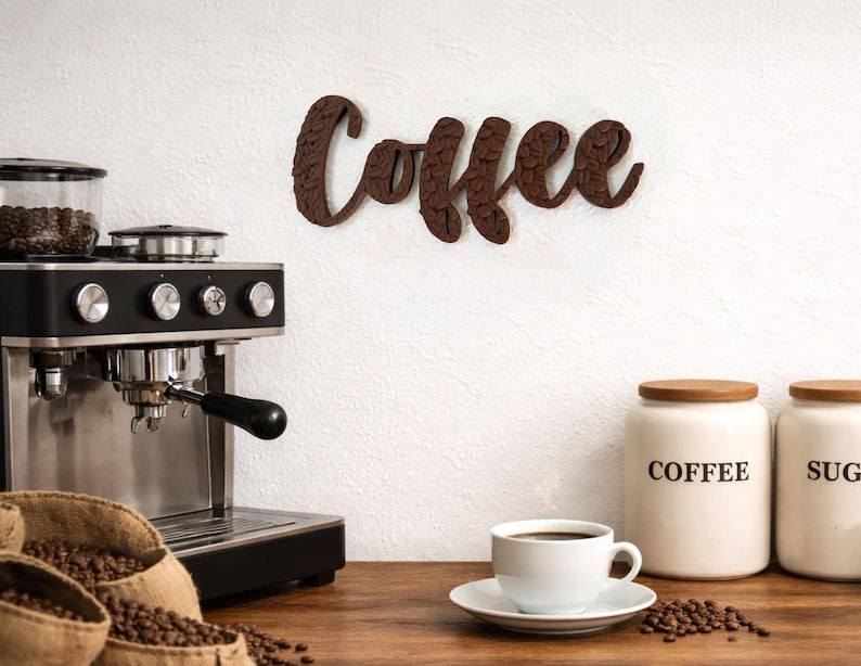 May include: A brown wooden "Coffee" sign hangs on a white wall above a coffee machine and a cup of coffee. The sign is in a cursive font with a textured design. Jars labeled "Coffee" and "Sugar" sit nearby, along with burlap sacks filled with coffee beans.