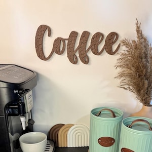 May include: A brown wooden sign that says "Coffee" in a cursive font, mounted on a white wall. The sign has a textured surface. A black espresso machine, a white cup, and two green canisters are in the foreground.