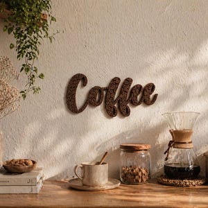 May include: A wooden sign spelling "Coffee" hangs on a white wall. A coffee setup includes a glass pour-over, a mug, a jar of beans, and a wooden grinder. A vase of dried flowers and books are on a wooden table.