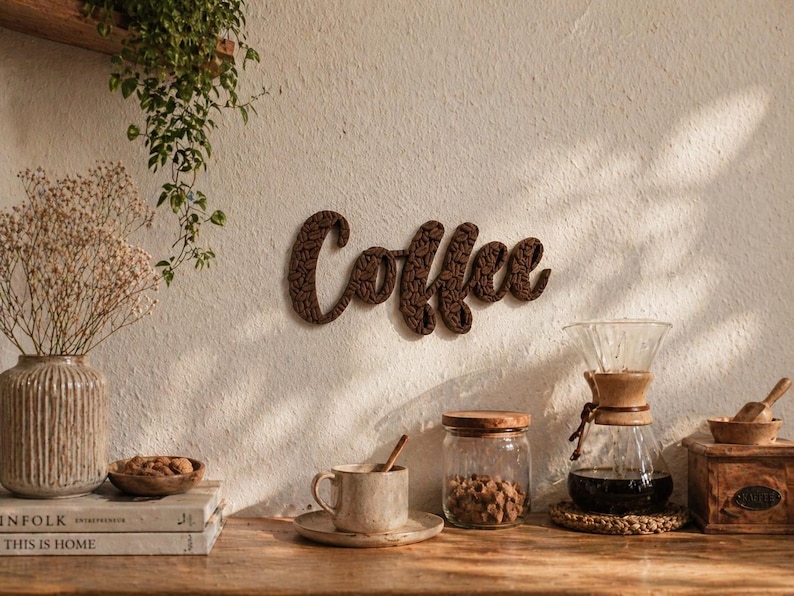 May include: A wooden sign spelling "Coffee" hangs on a white wall. A coffee setup includes a glass pour-over, a mug, a jar of beans, and a wooden grinder. A vase of dried flowers and books are on a wooden table.