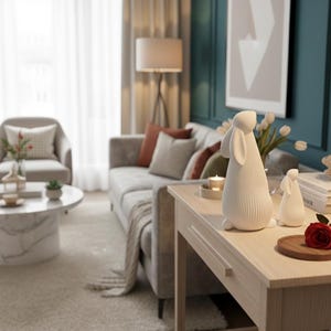 May include: A light wood side table displays two white ceramic rabbit figurines. A red rose sits on a small wooden plate. The room features a gray sofa, a marble coffee table, and a teal wall with a framed abstract art piece.