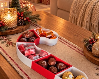 May include: A candy cane shaped serving dish divided into multiple red and white compartments. The dish is filled with various wrapped chocolates, including gold, red, and orange foil-wrapped candies, arranged in a festive holiday display.