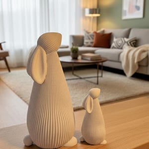 May include: Two white, textured rabbit figurines of different sizes. The larger rabbit has long ears and a tall body, while the smaller one is more compact. Both have a ribbed design and are placed on a wooden surface, suggesting a decorative theme.