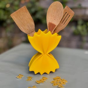 Könnte beinhalten: Ein leuchtend gelber Utensilienhalter in Form einer Farfalle-Nudelschleife, der drei hölzerne Küchenutensilien enthält, darunter einen Pfannenwender und einen Schlitzwender, mit mehreren kleinen trockenen Farfalle-Nudeln im Vordergrund.