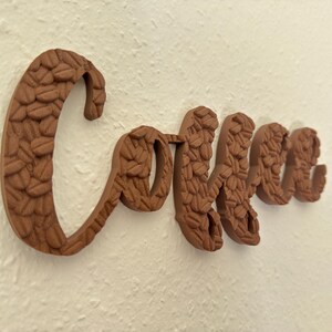 May include: A brown wooden sign spelling "Coffee" in a cursive font. The letters are textured with a pattern resembling coffee beans. The sign is mounted on a textured white wall, creating a warm, inviting aesthetic.