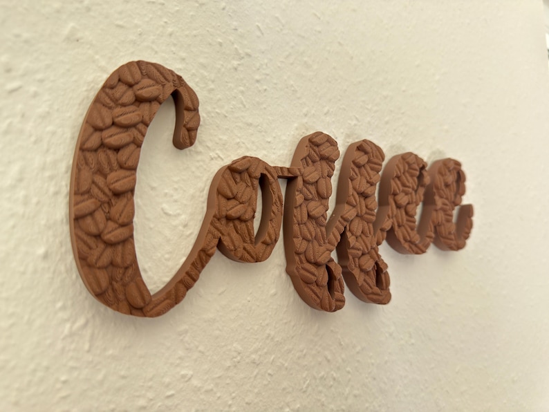 May include: A brown wooden sign spelling "Coffee" in a cursive font. The letters are textured with a pattern resembling coffee beans. The sign is mounted on a textured white wall, creating a warm, inviting aesthetic.
