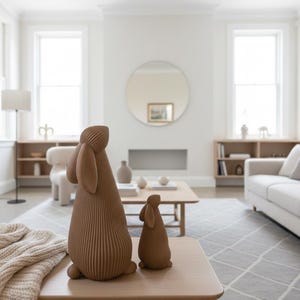 May include: Two brown rabbit sculptures with textured, vertical lines sit on a light wood table. The larger rabbit has long ears. The background shows a living room with a neutral color palette, a sofa, and a geometric rug.