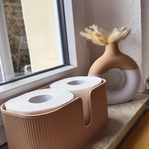 May include: A light peach-colored rectangular storage container with vertical ribbed detailing holding two rolls of white toilet paper, positioned next to a two-toned beige and white circular ceramic vase containing dried tan grass stems.