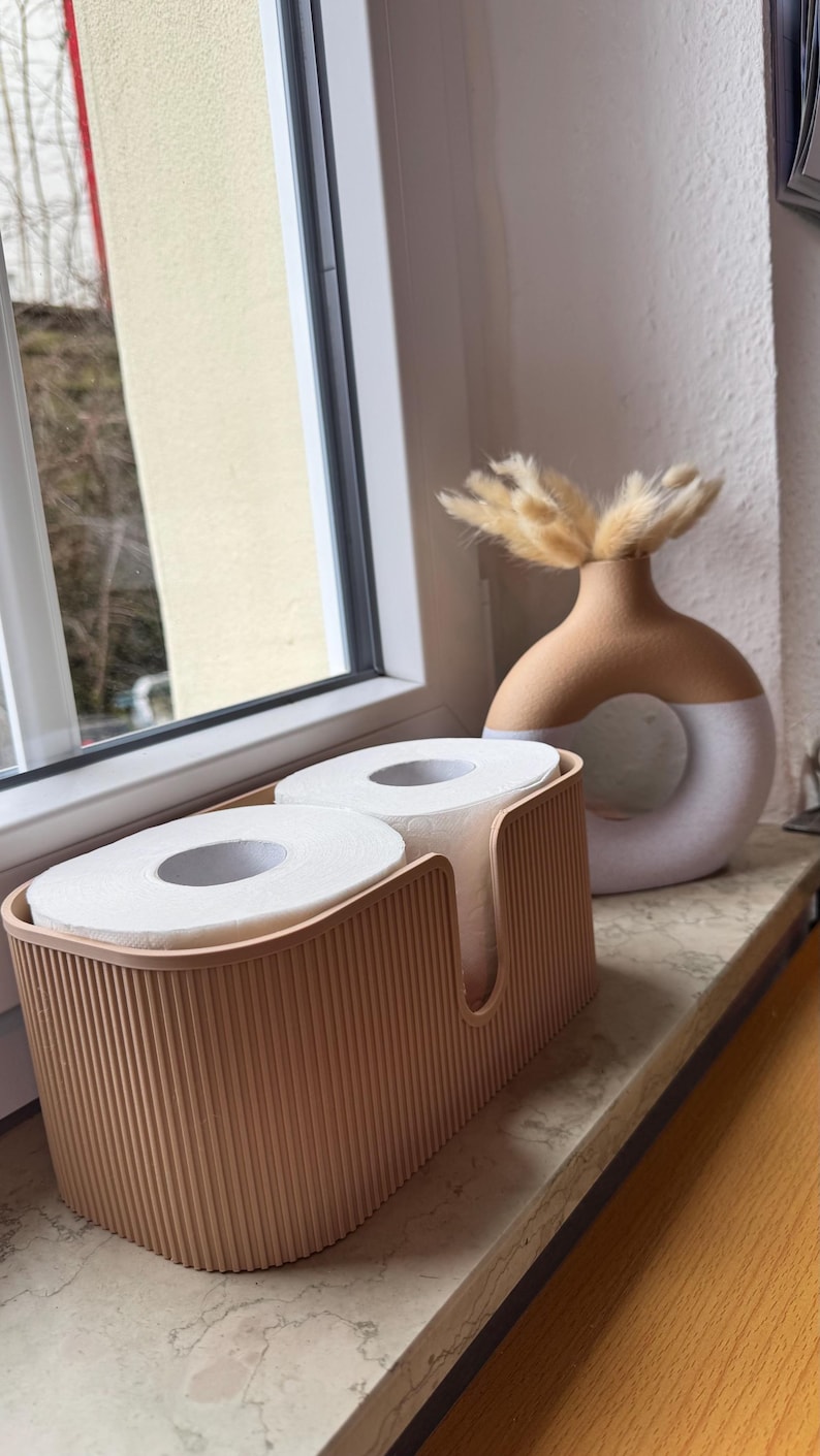 May include: A light peach-colored rectangular storage container with vertical ribbed detailing holding two rolls of white toilet paper, positioned next to a two-toned beige and white circular ceramic vase containing dried tan grass stems.