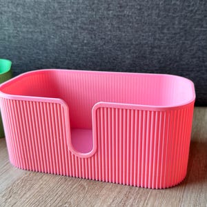 May include: A bright pink rectangular storage container with a ribbed, vertical texture on the exterior walls. The container features a U-shaped cutout on the front panel, providing an open access point to the interior space of the storage bin.