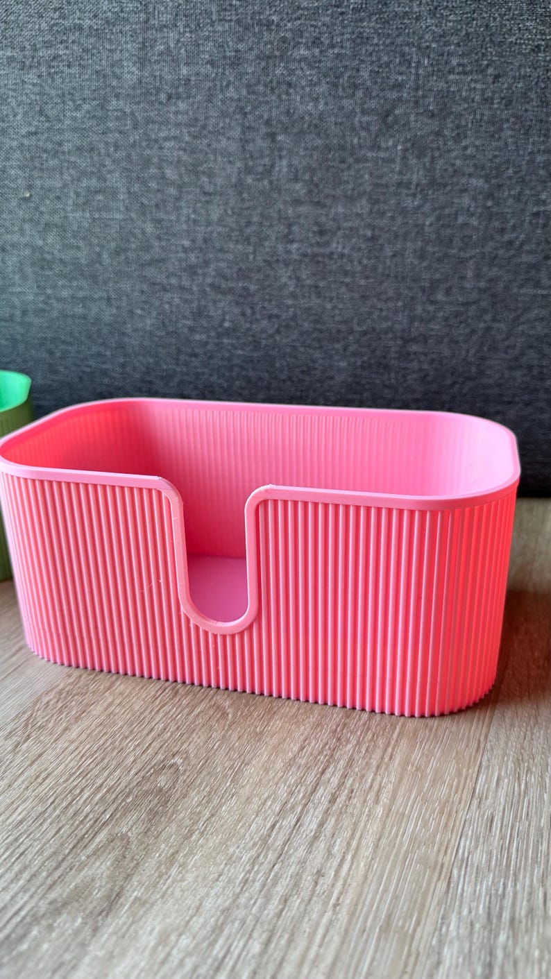 May include: A bright pink rectangular storage container with a ribbed, vertical texture on the exterior walls. The container features a U-shaped cutout on the front panel, providing an open access point to the interior space of the storage bin.