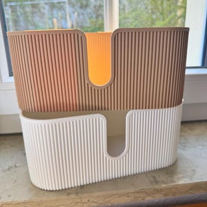 May include: A two-tiered storage organizer featuring a ribbed texture on the exterior. The top section is a tan color, while the bottom section is white. Both tiers have a U-shaped cutout on the front panel, allowing for easy access to items stored inside.