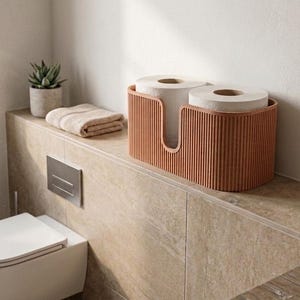 May include: An oval-shaped, terracotta-colored storage bin with a vertical ribbed texture and a U-shaped cutout on the front. The container holds two white toilet paper rolls, featuring a rounded design that accommodates the rolls side by side.