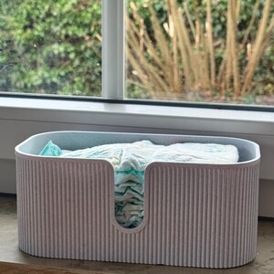May include: A light gray, oval-shaped storage bin with a vertical ribbed texture and a U-shaped cutout on the front. The bin is filled with a stack of folded, light blue and white patterned disposable diapers, visible through the front opening.