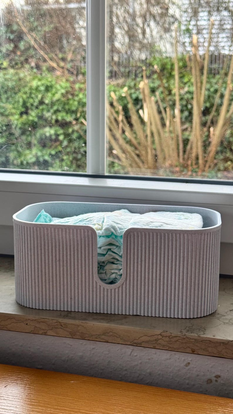 May include: A light gray, oval-shaped storage bin with a vertical ribbed texture and a U-shaped cutout on the front. The bin is filled with a stack of folded, light blue and white patterned disposable diapers, visible through the front opening.