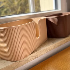 May include: Two rectangular storage bins with a vertical ribbed texture. The front bin is light beige and features a U-shaped cutout on the front panel. The bin positioned behind it is a darker brown color. Both containers have rounded corners.