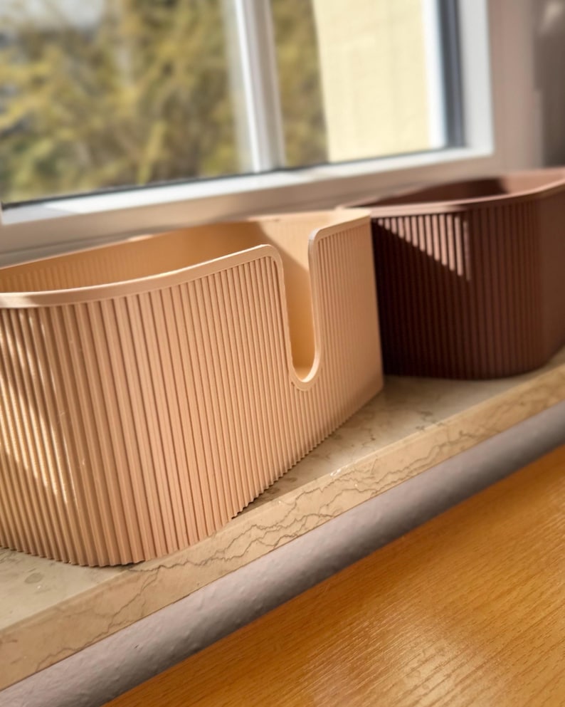 May include: Two rectangular storage bins with a vertical ribbed texture. The front bin is light beige and features a U-shaped cutout on the front panel. The bin positioned behind it is a darker brown color. Both containers have rounded corners.