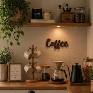 May include: A coffee station with a wooden shelf holding coffee-related items. Includes a coffee grinder, jars of beans, and a wooden sign that says "Coffee". A wooden mug tree holds six mugs. A coffee maker and kettle are on the counter.