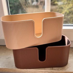 May include: Two oval-shaped storage bins with a vertical ribbed texture on the exterior. The top bin is light beige, and the bottom bin is dark brown. Each bin features a U-shaped cutout on the front side, allowing for easy access to contents.