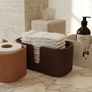 May include: Three rectangular storage bins with a vertical ribbed texture. The left bin is terracotta, the middle is dark brown holding folded white cloths, and the right is cream, containing a dark brown bottle, a silver bottle, and a small jar.