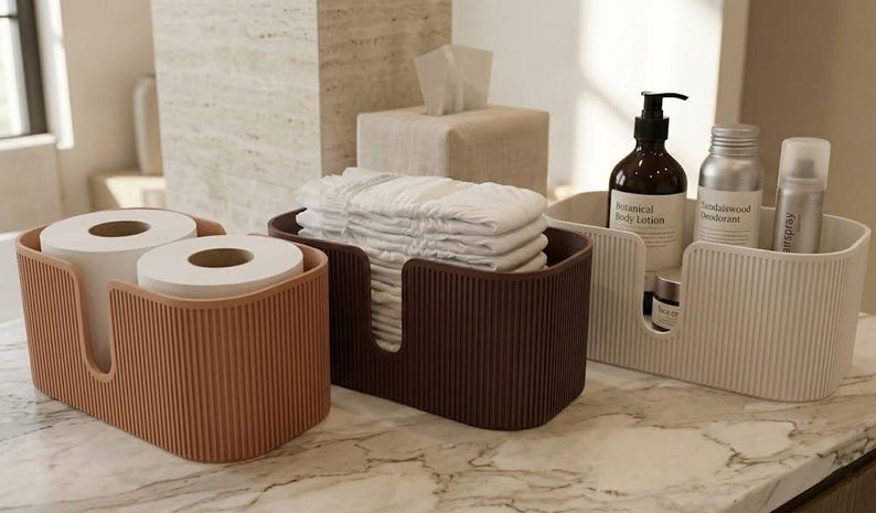 May include: Three rectangular storage bins with a vertical ribbed texture. The left bin is terracotta, the middle is dark brown holding folded white cloths, and the right is cream, containing a dark brown bottle, a silver bottle, and a small jar.