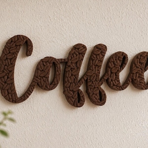 May include: A brown wooden sign spelling "Coffee" in a cursive font. The letters are filled with a pattern of coffee beans. The sign is mounted on a textured, off-white wall, with a hint of greenery in the lower left corner.