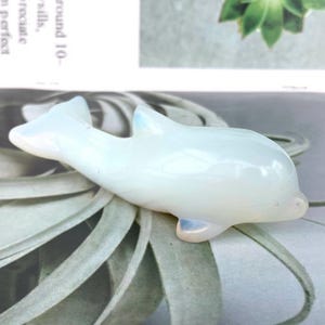 May include: A translucent, opalescent dolphin ornament. The sculpture has a smooth, polished surface with subtle blue highlights on the fins and tail. The dolphin is lying down, with its body curved in a graceful pose.