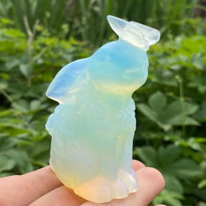 May include: A translucent, opalescent bunny figurine with a light blue and yellow color gradient. The bunny is carved with detailed features, including ears and a small tail. The figurine is held in a hand against a backdrop of green foliage.