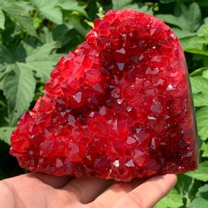 May include: A vibrant, heart-shaped cluster of deep red crystals. The crystals are densely packed, with sharp, geometric facets catching the light. The overall form is rough and natural, with a rich, saturated color. The background features green foliage.