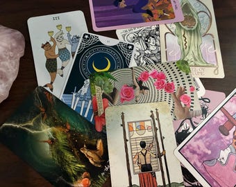 Könnte beinhalten: Eine Sammlung von Tarotkarten mit verschiedenen Illustrationen, darunter Figuren, Symbole und Szenen. Die Karten weisen eine Reihe von Farben und künstlerischen Stilen auf, wobei einige Karten Text und Zahlen anzeigen. Die Karten sind auf einer dunklen Oberfläche angeordnet.