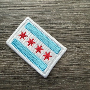 May include: A rectangular embroidered patch featuring the Chicago flag design. The patch has a white background with two horizontal light blue stripes and four bright red six-pointed stars centered between the stripes, all bordered by white stitching.