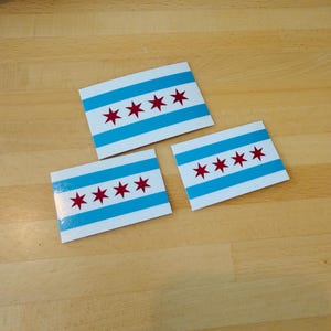 Puede incluir: Tres pegatinas rectangulares con el diseño de la bandera de Chicago. Cada pegatina tiene un fondo blanco con dos franjas horizontales azul claro y cuatro estrellas rojas de seis puntas. Las pegatinas están sobre una superficie de madera marrón claro.
