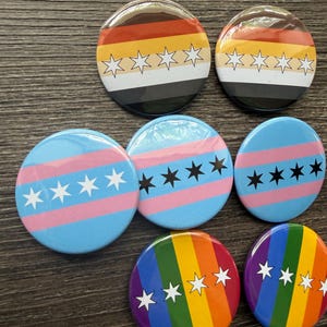 May include: Seven circular pin-back buttons featuring various flag designs. Two buttons display horizontal stripes of brown, orange, yellow, white, and black with white stars. Three buttons show horizontal stripes of blue and pink with stars. Two buttons feature vertical rainbow stripes with white stars.