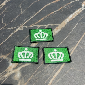 May include: Three green and white embroidered patches. Each patch is rectangular with a green background and a white crown design. The patches have a black border and are displayed on a marble surface.