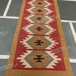 Puede incluir: Una alfombra rectangular con un patrón geométrico. La alfombra presenta un campo central rojo con formas de diamantes repetidas en blanco, gris y negro. El borde es de color marrón claro. La alfombra está hecha de un material tejido.