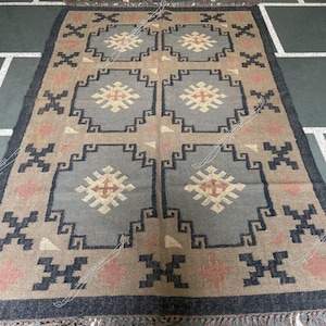 May include: Rectangular jute rug with a geometric pattern. The rug features a central design of repeating squares in shades of gray, beige, and pink, outlined with dark blue shapes. The rug has a dark blue border and fringe.