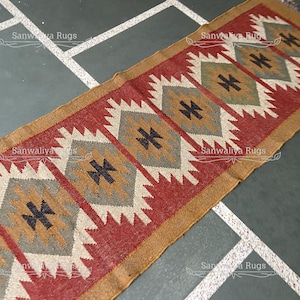 Puede incluir: Una alfombra de corredor rectangular con un patrón geométrico. La alfombra presenta un fondo rojo con formas de diamante en blanco, gris y beige, acentuadas con cruces negras. El borde es beige.