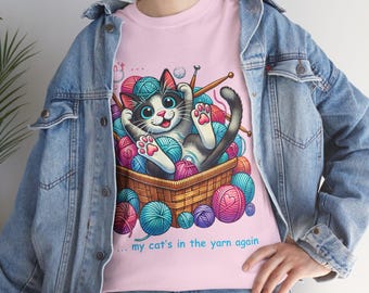 May include: Pink t-shirt featuring a cartoon cat in a basket of yarn. The cat has a black and white coat, blue eyes, and pink paws. The text on the shirt reads "... my cat's in the yarn again."