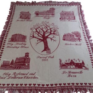 May include: A cream-colored throw blanket with a burgundy design featuring illustrations of buildings and a tree. The blanket has a fringed edge and a border of burgundy hearts. Text includes "Sacred Oak 1993" and building names.