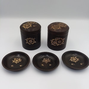 May include: A set of dark brown, lacquered tea canisters and saucers. The canisters are cylindrical with lids, and the saucers are round. Each piece is decorated with a gold floral design.