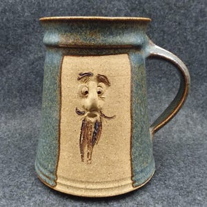 May include: A handmade ceramic mug with a sculpted face design. The mug features a tan face with a long beard, framed by a blue-green background. The handle is brown, and the mug has a rustic, artisanal appearance.