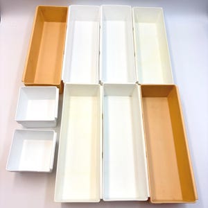 May include: A collection of eight rectangular plastic storage bins in varying sizes. The set includes four long, narrow bins in white and light orange, and four smaller, square-shaped white bins, all featuring open tops and smooth walls.