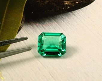 Könnte beinhalten: Ein lebendiger, smaragdgeschliffener grüner Edelstein. Der Edelstein ist rechteckig mit facettierten Kanten, die das Licht reflektieren. Der Stein liegt auf einer hellen Oberfläche, mit einem dunklen Felsen und einem Blatt im Hintergrund.